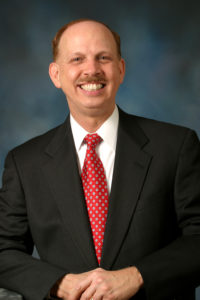 Author Headshot in dark grey suit and red tie with arms folded smiling in front of dark background
