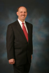 Author headshot standing in dark suit with red tie against a dark grey background